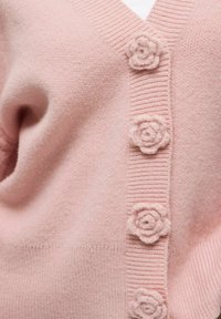 Roze cardigan met een gestructureerde brei, V-hals en decoratieve bloemenknopen langs de voorkant, die de zachte, warme stof benadrukken.