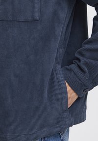 Tengerészkék kordbársony dzseki bordázott textúrával, egyetlen mellzsebbel, gombbal záródó mandzsetta részekkel és a side seam-ekbe integrált zsebekkel.