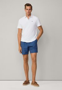 Bijela polo majica, plavi kratke hlače s geometrijskim uzorkom i smeđe espadrile. Model stoji s jednom rukom u džepu.