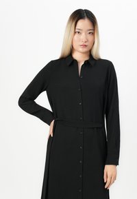 Black long-sleeve dress with a collared neck, button front, and a tied waist. Made of textured fabric, offering a tailored yet relaxed fit.