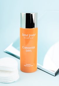 Lys orange flaske med Vitamin C Brightening Tonic med en sølvhætte, placeret ved siden af vatrondeller og en spejlflade mod en blå baggrund.