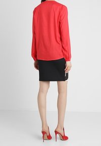 Donna che indossa una blusa lunga rossa a maniche, una gonna nera con dettagli floreali laterali e scarpe rosse con tacco alto, in piedi davanti a uno sfondo bianco.