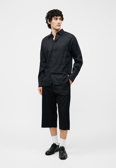 Άνδρας με σγουρά μαλλιά φοράει μαύρο πουκάμισο με μοτίβο μακρυμάνικο, μαύρο φαρδύ παντελόνι καρό, λευκές κάλτσες και μαύρα παπούτσια, στέκεται με το χέρι στην τσέπη.