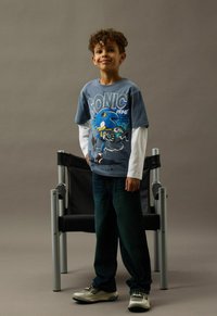 Grå T-shirt med ett Sonic the Hedgehog-tryck över en vit långärmad tröja, matchad med mörka jeans och ljusa sneakers.