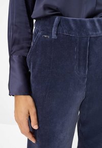 Marineblauwe corduroy broek met verticale ribbelstructuur, voorzien van een platte tailleband, zijzakken en een klein merklabel op de tailleband.