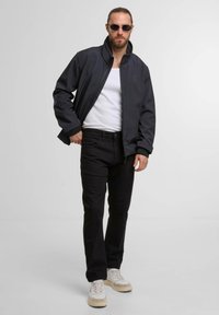 Black zippered jacket with a high collar, white tank top, black pants, and beige sneakers. Round sunglasses complete the look.