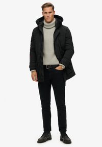 Chaqueta de plumas negra con capucha, suéter de cuello alto gris de punto, pantalones negros y botas negras. Tiene bolsillos laterales y una textura suave.