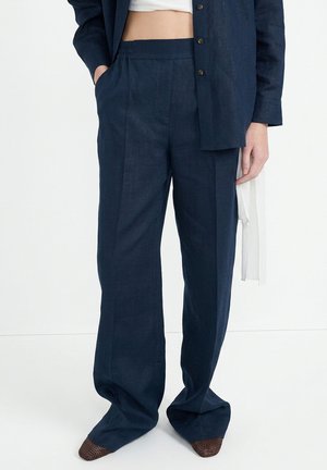 Persoon die loszittende donkerblauwe broek met elastische taille en bijpassend donkerblauw overhemd met knopen draagt, staand met één hand in de zak.