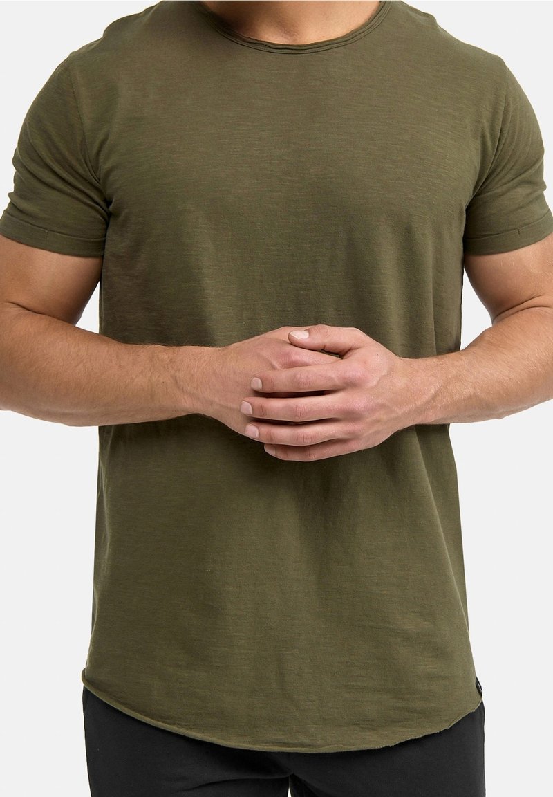 Camiseta de manga corta color verde oliva en tejido suave y texturizado, con cuello redondo y dobladillo redondeado, llevada con las manos entrelazadas frente al cuerpo.