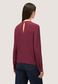 Blusa bordeaux con maniche lunghe, dettaglio a occhiello sulla schiena, texture liscia e bottoni sugli polsini. Abbinata a jeans blu scuro.