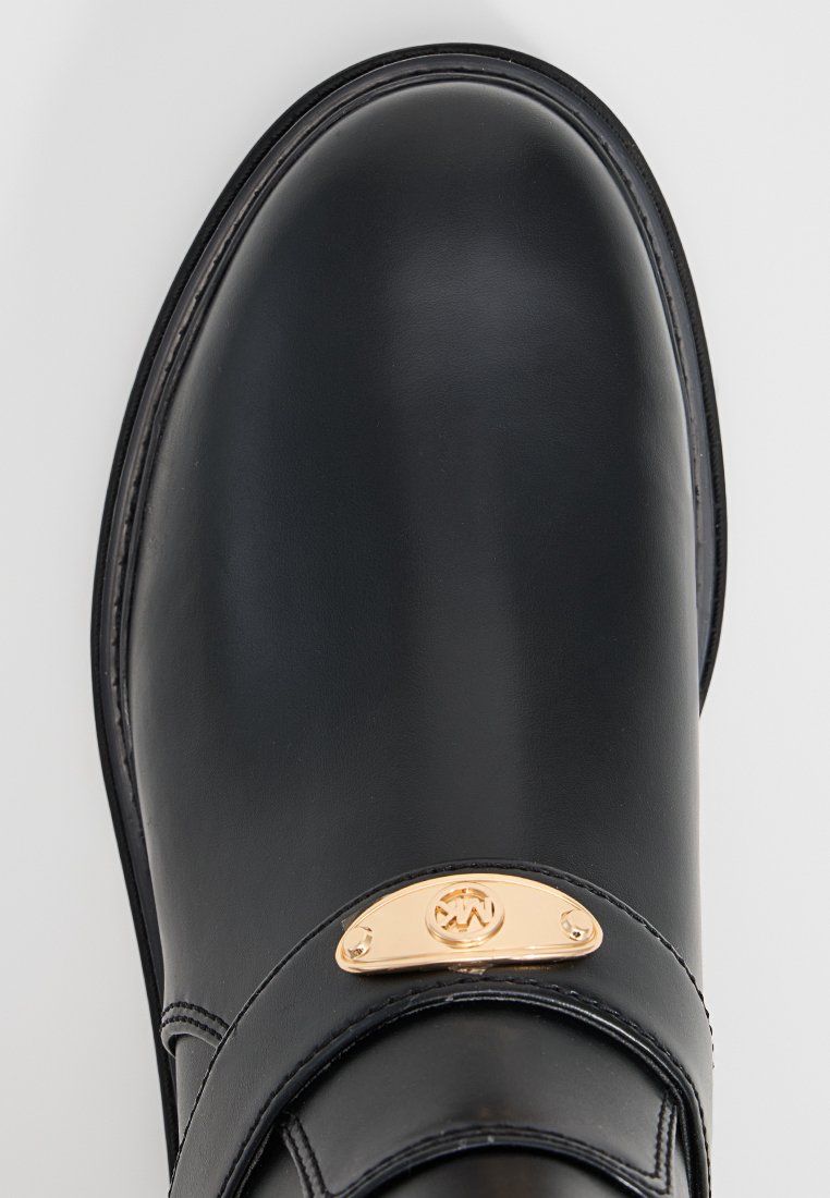 Czarny skórzany loafers z zaokrąglonym czubkiem i złotą metalową plakietką logo na wierzchu, widocznym przeszyciem wzdłuż krawędzi podeszwy.