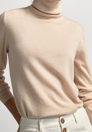 Pull à col roulé beige à manches longues associé à un pantalon blanc taille haute avec de gros boutons marron.