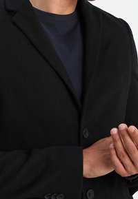 Zwarte blazer met gelapte revers, enkelzijdig ontwerp en twee knopen aan de voorkant. Draagt over een marineblauw T-shirt met ronde hals. Soepele stoftextuur.