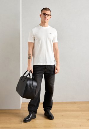 Hvit kortermet t-skjorte, svarte jeans, svart skinnsekk med håndtak. Brukeren har kort hår og briller, står på tregulv.
