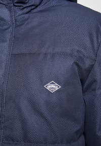 Marinblå jacka med ribbad struktur, med en liten vit diamantlogotyp märkt "ORIGINALS" på vänster sida.