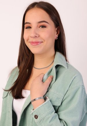 Zilveren choker ketting en een gestructureerde grijze armband gedragen met een lichtgroen corduroy shirt. Huidteint is licht, haar is bruin en steil.