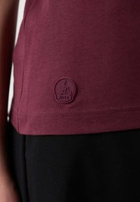 Camiseta burdeos hecha de una tela suave, con un logo redondo púrpura que presenta un personaje en bicicleta en el dobladillo. Pantalones negros debajo de la camiseta.