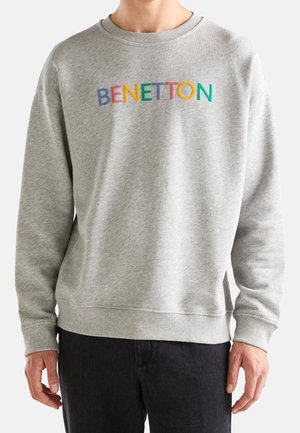 Osoba ubrana w jasnoszary sweter z wielokolorowym napisem "BENETTON" na piersi oraz czarne spodnie na jednolitym tle.