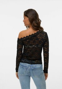Zwarte kanten top met een off-the-shoulder ontwerp, lange mouwen en golvende randen. Draagt bij lichte blauwe spijkerbroeken, met zichtbaar textuur en patroon.