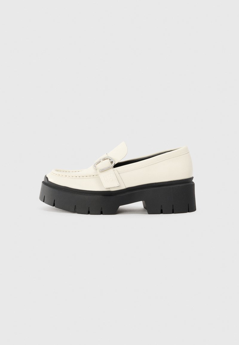 Valkoinen nahkainen loafers-kengät, joissa on paksu musta pohja, esillä suorakaiteen muotoinen hopeinen solki ja näkyvät ompelusaumat päällä.