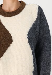 Pull en tricot crème, marron et gris foncé, présentant un design en blocs de couleur avec des sections diagonales et une finition texturée. Col rond.