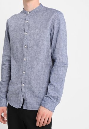 Homme portant une chemise à manches longues bleu clair avec col officier et pantalons noirs, sur un fond uni.