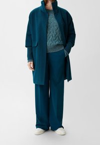 Person in türkisfarbener, weit geschnittener Hose, türkisfarbener, strukturiertem Pullover, passendem türkisfarbenem Mantel mit Taschen und weißen Sneakers, steht vor einem neutralen Hintergrund.