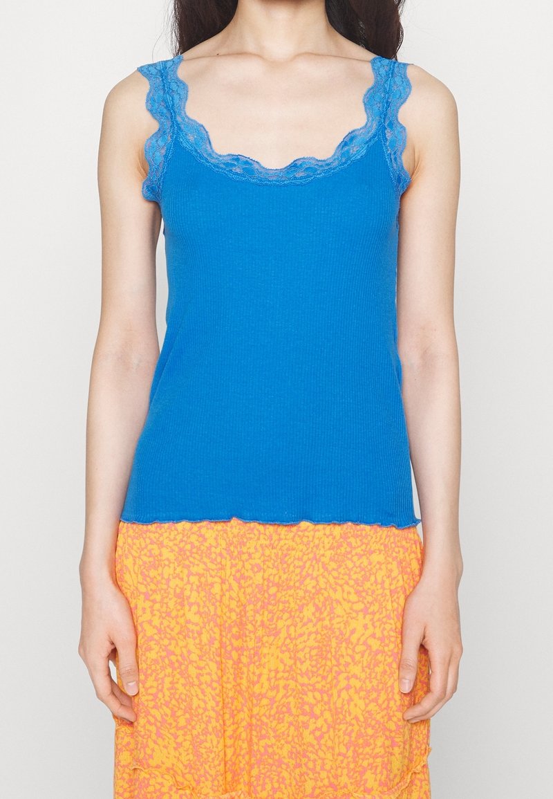 Débardeur ribbed bleu avec dentelle, présentant un décolleté et des bretelles à bords festonnés, associé à une jupe orange à motifs.