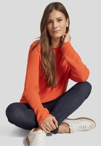 Oranges Strickpullover mit rundem Ausschnitt, kombiniert mit dunkelblauen Jeans und weißen Sneakers; glatte Textur und lässiges Design.