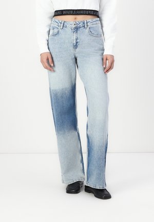 Model iført to-tonede lyse og mellemblå vidde jeans, sorte sko med firkantet tå og en hvid cropped top med synlig sort taljebåndstekst.