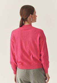 Pull en tricot rose avec un col rond, des poignets et un ourlet côtelés. Texture lisse, coupe décontractée, et manches longues présentées de dos.