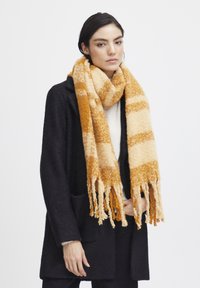 Écharpe texturée avec des rayures alternées de jaune chaud et de crème, dotée de bords frangés, portée sur un manteau sombre.