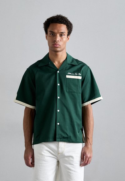 Camicia verde a maniche corte con bottone frontale, polsini beige a contrasto e una singola tasca sul petto con testo bianco ricamato.