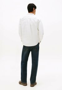 Person standing with back facing camera, wearing a white long-sleeve shirt, dark blue jeans, and brown hiking shoes on a plain background.
