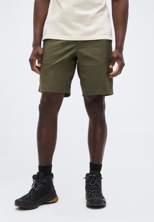 Mees kannab oliivrohelisi lühikesi pükse, musti matkasaapaid mustade sokkidega, seisab ühevärvilise tausta ees.