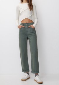 Femme portant un top blanc à manches longues crop, un pantalon vert taille haute retroussé, des chaussettes grises et des baskets blanches avec des semelles en gomme.