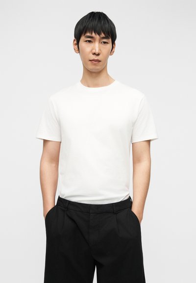 Νεαρός άνδρας με κοντά μαύρα μαλλιά, που φοράει απλό λευκό μπλουζάκι και μαύρο παντελόνι, με τα χέρια στις τσέπες, στέκεται μπροστά σε ανοιχτόχρωμο φόντο.