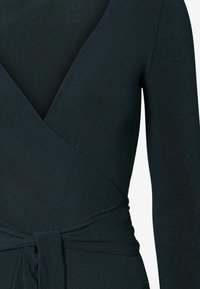 Top avvolgente verde scuro con maniche lunghe, realizzato in un tessuto liscio e elastico. Presenta un dettaglio con fiocco in vita e un design a scollo a V.