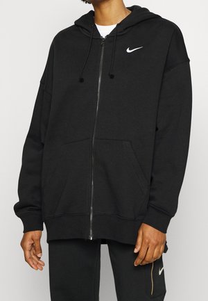 Personne portant un sweat à capuche zippé noir Nike avec poches avant et un pantalon noir assorti avec une poche zippée latérale et le logo Nike.