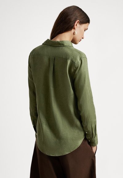 Zaļa pogājama krekla izgatavots no teksturētas auduma, ar apkakli, garām piedurknēm un noapaļotu apakšu. Redzams aizmugurējais krokas detaļas.