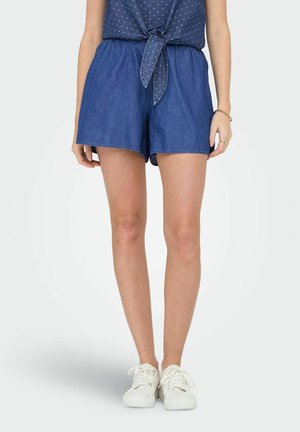 Shorts in denim con vita elasticizzata, di colore blu navy e orlo sfrangiato. Abbinati a sneakers bianche e una maglietta a pois con nodo davanti.