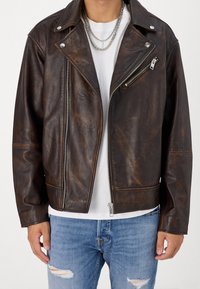 Ruskea keinonahkainen moottoripyörätakki hopeisilla vetoketjuyksityiskohdilla, kaulus ja rintatasku. Päällä valkoinen paita ja kulutetut farkut.