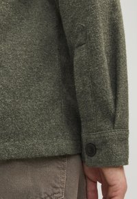 Manica di giacca grigia in feltro caratterizzata da un bottone nero al polsino, con una texture liscia e una vestibilità sagomata. Dettaglio delle cuciture visibile lungo il braccio.