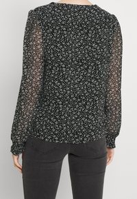 Svart blus med vita prickar, genomskinliga långa ärmar, rynkade manschetter och rund halsringning. Bakifrån syns det blommönster och skärning.