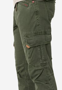 Cargo-Hosen in Olivgrün, aus einem strukturierten Stoff, mit seitlichen Taschen und dezenten Stichdetails. Inklusive eines roten Akzents am Reißverschluss.