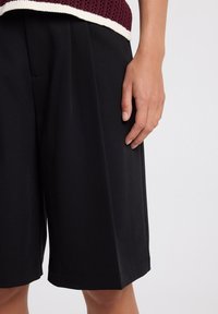 Black tailored shorts with side pockets, featuring a pleated design and smooth texture. A hand lightly rests on the fabric.
