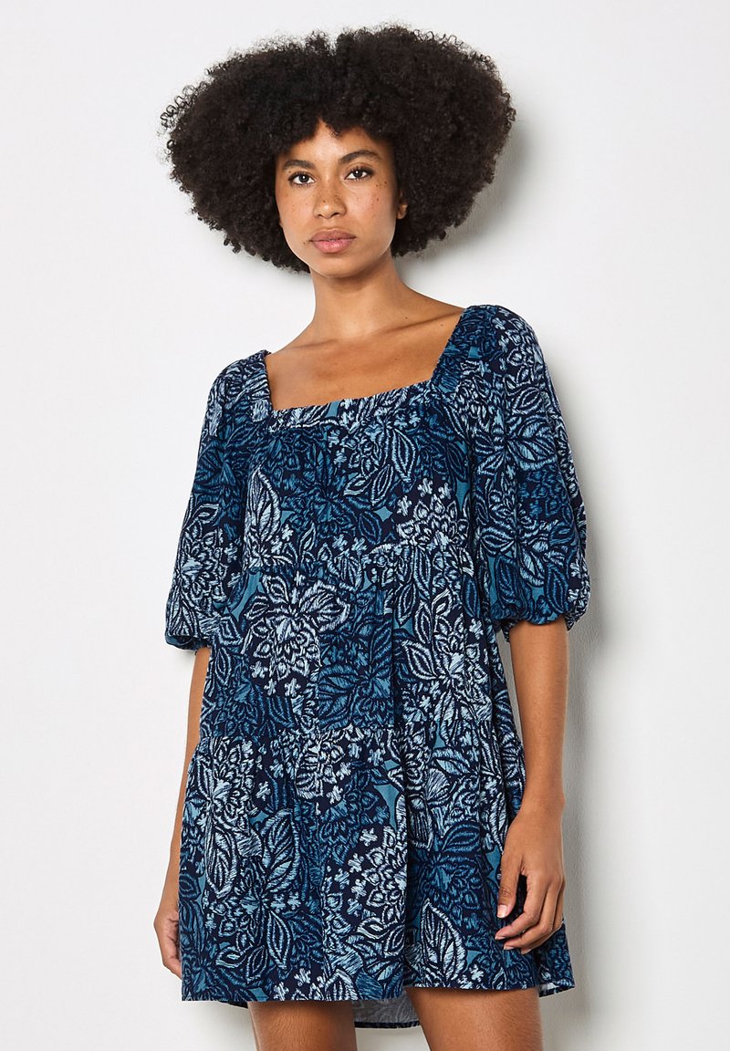 Femme aux cheveux afro bouclés portant une robe bleue à motif floral avec des manches bouffantes et un décolleté carré, debout devant un fond uni.