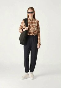 Brown plaid shirt, black elastic waistband trousers, white sneakers, and black shoulder bag. Sunglasses and minimal accessories.