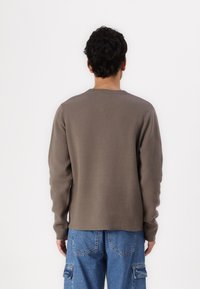 Brauner gestrickter Pullover mit runden Halsausschnitt und langen Ärmeln, kombiniert mit hellblauen Cargo-Hosen. Der Stoff wirkt strukturiert und weich.