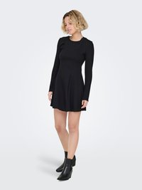 Robe noire à manches longues avec un corsage ajusté, une jupe évasée et une texture lisse. Associée à des bottines noires à bout pointu et talon compensé.